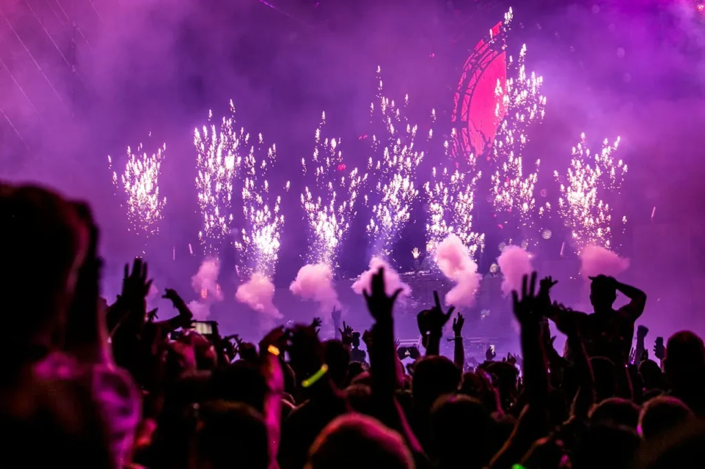 Estopins de fogos acesos no palco em primeiro plano; público e participantes ao fundo.